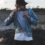 Man in a denim jacket and cap on a rocky outdoors setting, showcasing casual fashion.
