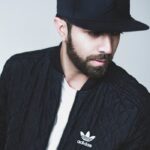 Portrait of a man in a black Adidas jacket and cap, looking down.