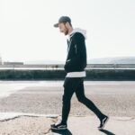 Casual male in a cap and hoodie walks along a sunny urban street.